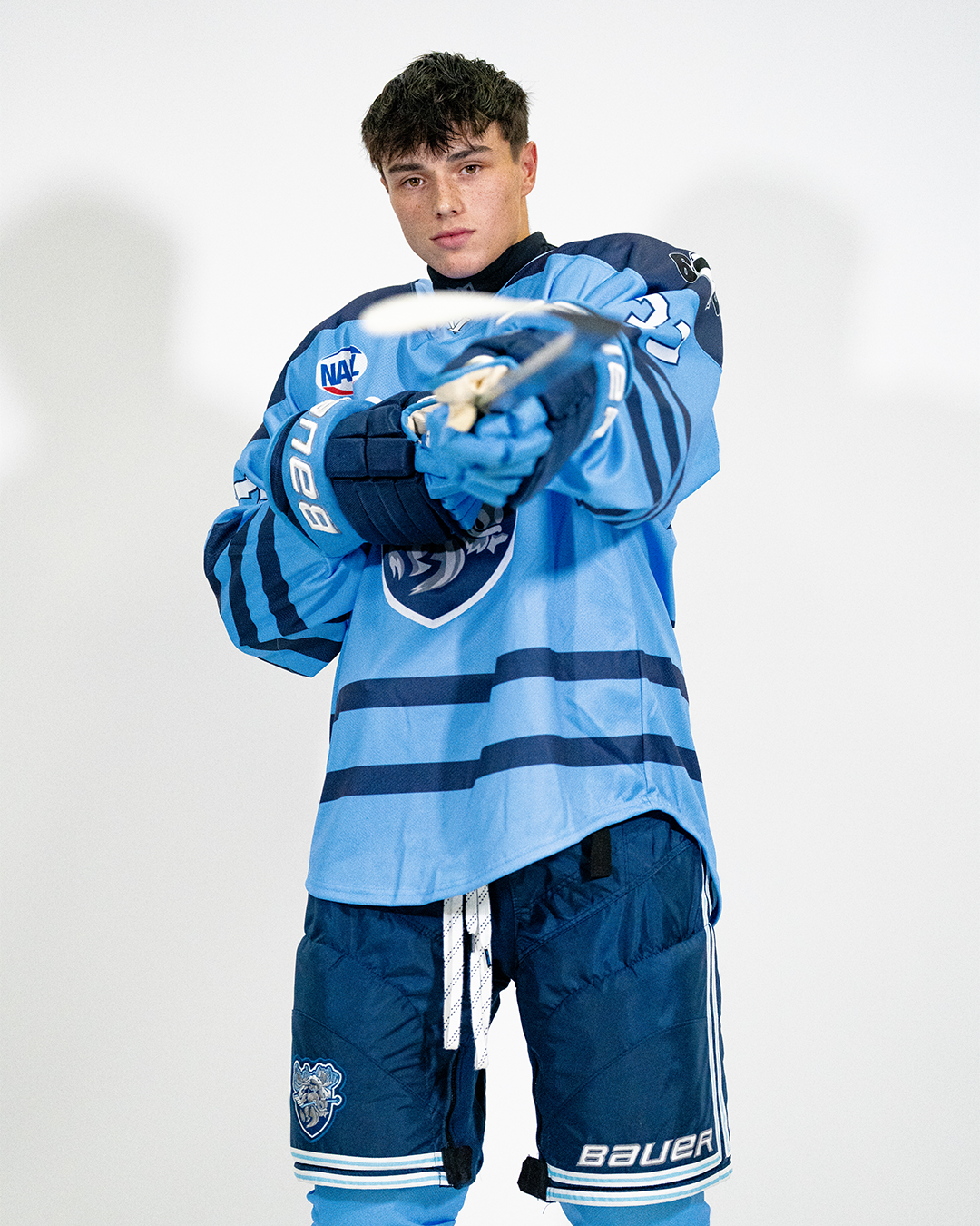 A young male hockey player wearing a blue jersey and shorts, holding a hockey stick, standing against a plain white background.