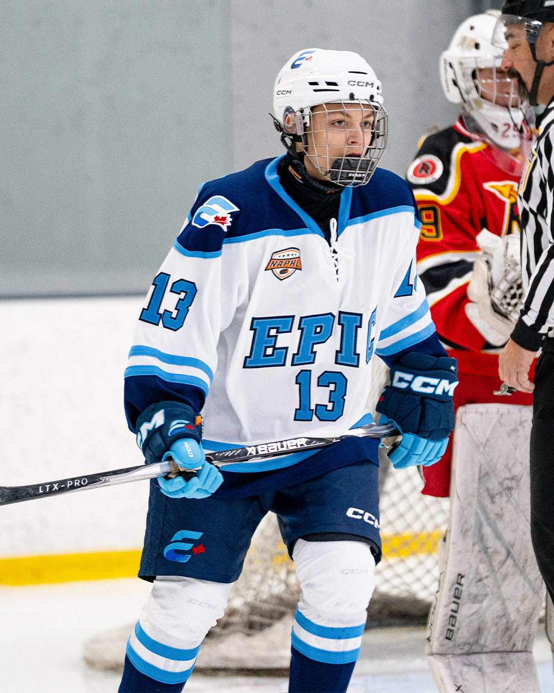 An ice hockey player in a white and blue uniform with the number 13 on his sleeve, holding a hockey stick, standing on the ice rink near the goal, wearing a helmet with a cage face mask. Another player in red and black gear and a referee are in the background.