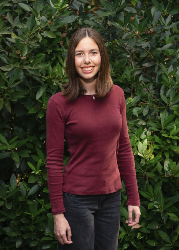 A young woman with shoulder-length brown hair, wearing a maroon long-sleeve shirt and dark jeans, standing outdoors in front of green leafy bushes, smiling at the camera.