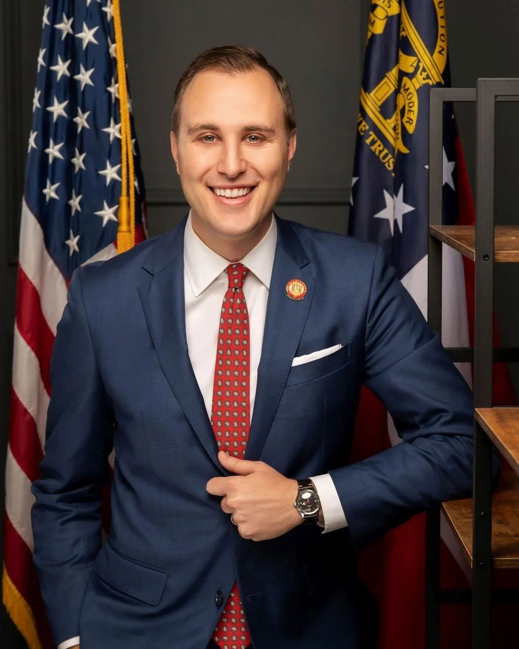 A man in a blue suit and red patterned tie smiling for the camera. He is standing in front of an American flag and a government flag, with a dark background and a wooden shelf visible to his right.