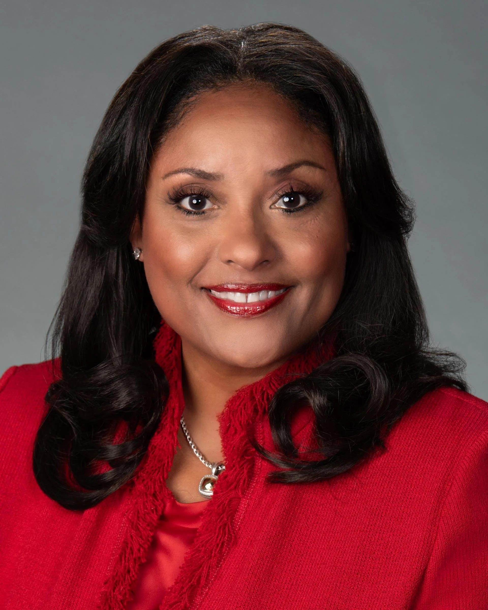 Close-up of a woman with dark hair, wearing a red sweater and a silver heart-shaped necklace, smiling.