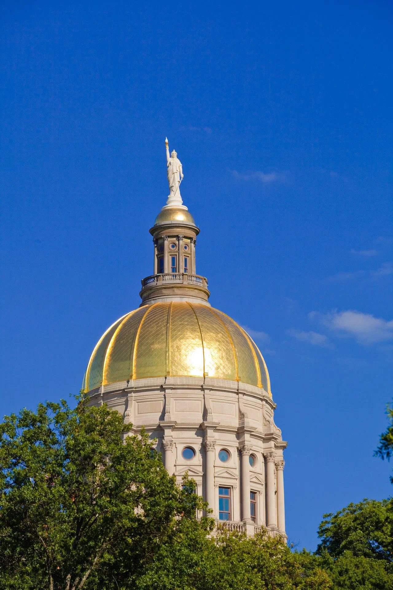 Capitol building with a golden dome and a statue on top, against a clear blue sky with some trees in the foreground.