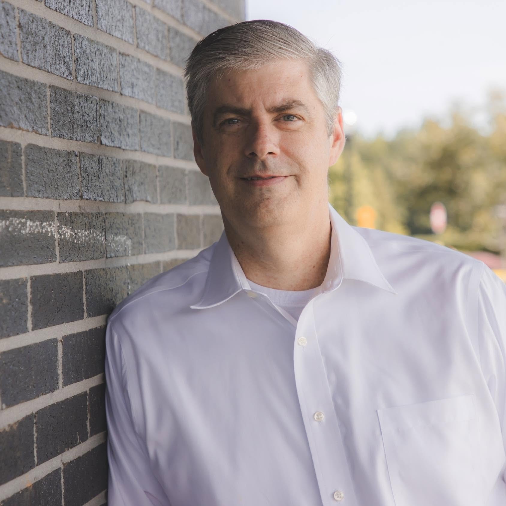 A middle-aged man with short, greying hair and blue eyes, wearing a white dress shirt, leaning against a brick wall outside with blurred trees and sky in the background.