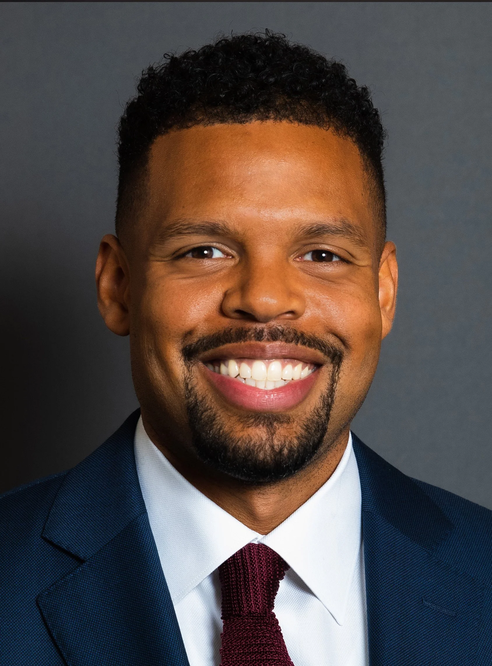 Close-up of a smiling man wearing a navy blue suit, white shirt, and burgundy tie, with a well-groomed beard and curly hair, against a dark gray background.