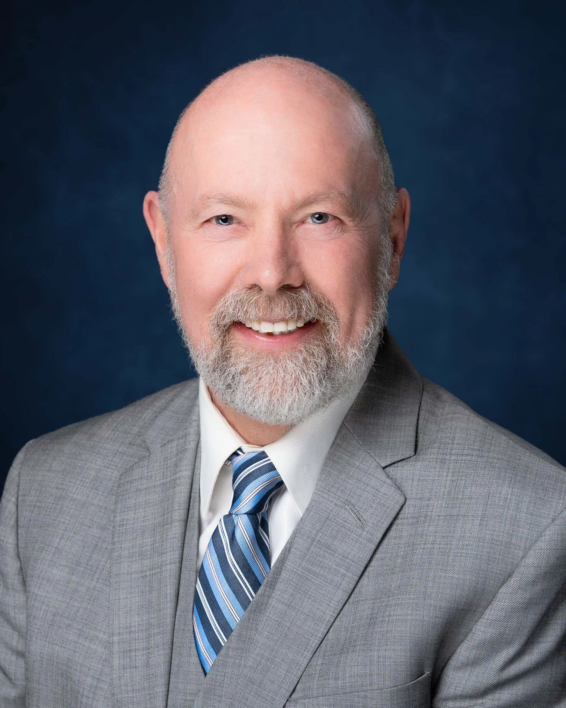 Close-up of a smiling older man wearing a gray suit, white shirt, and striped blue tie, with a dark blue background.