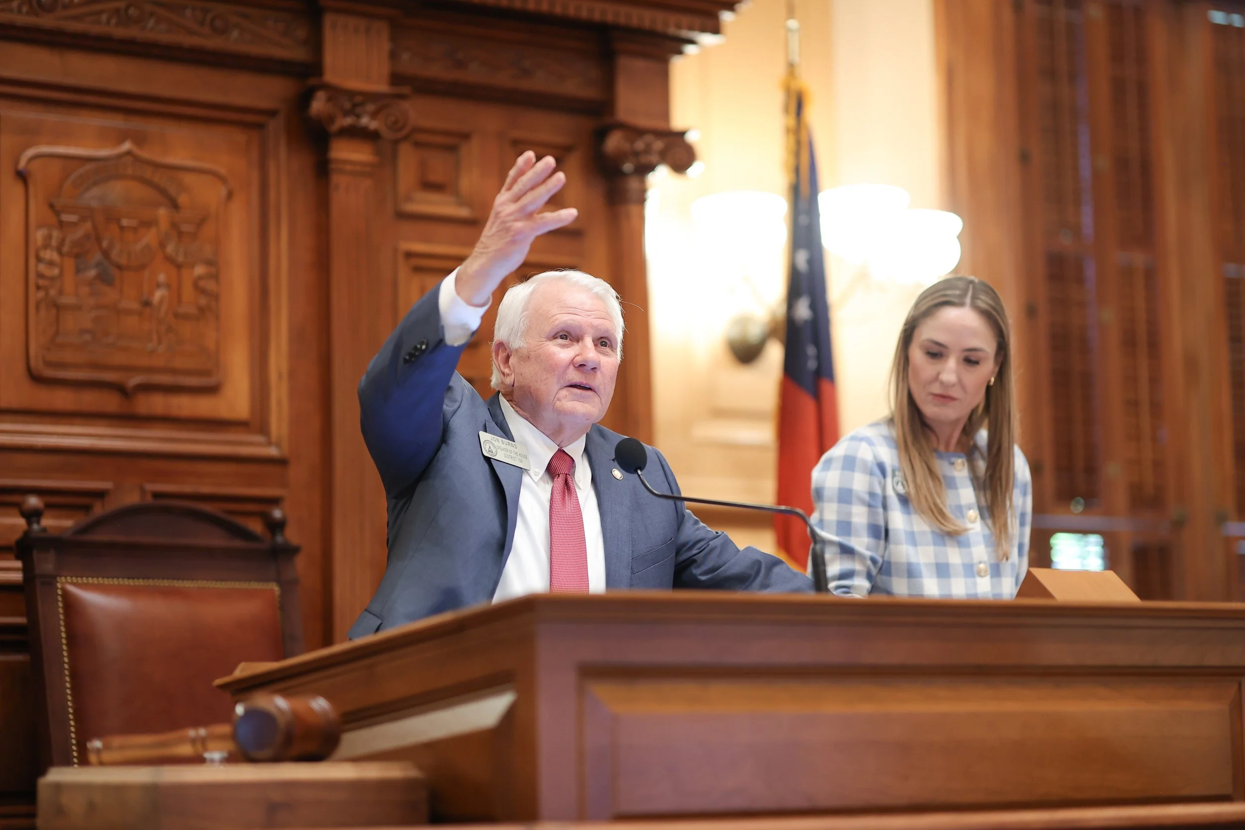 An elderly man in a suit with a red tie is speaking at a formal event, raising his hand. A young woman in a checkered shirt is sitting beside him, looking down. They are in a wood-paneled room with a flag in the background.