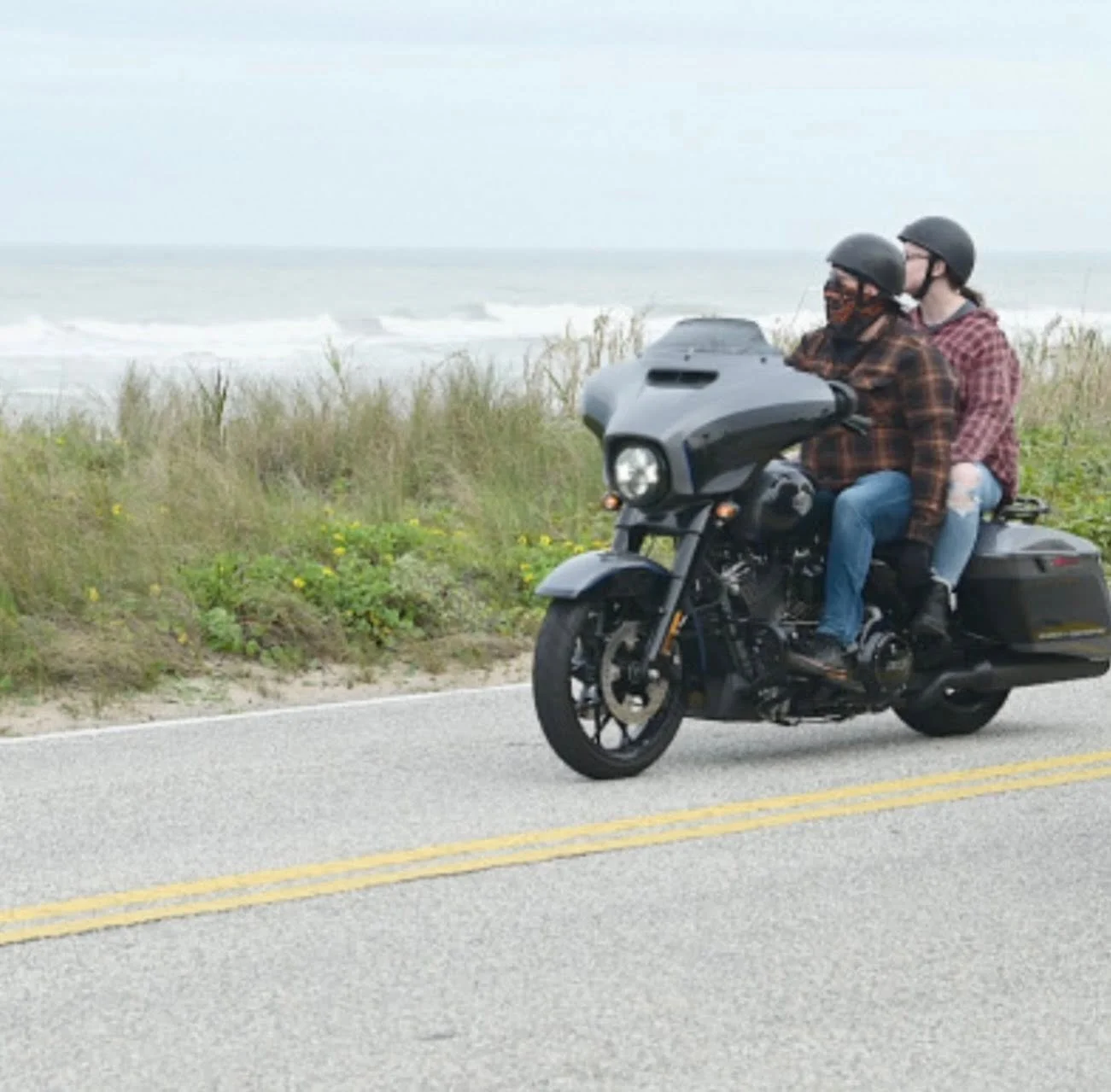 Two people riding a black motorcycle along a coastal road with grassy dunes and the ocean in the background. Both are wearing helmets, and the passenger is wearing ripped jeans.
