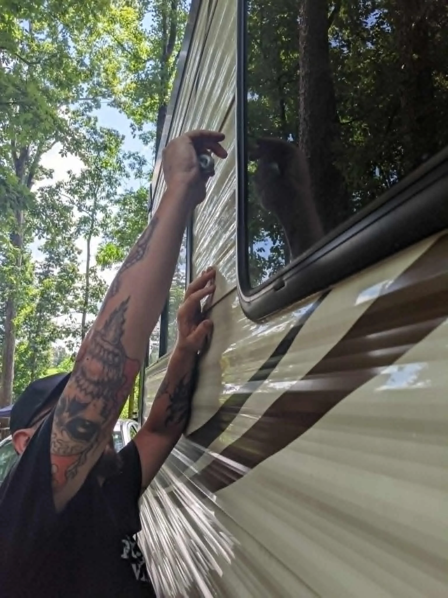 Person with tattooed arms touching or cleaning the side of a beige vehicle with reflective surface, outdoors with trees and blue sky in the background.