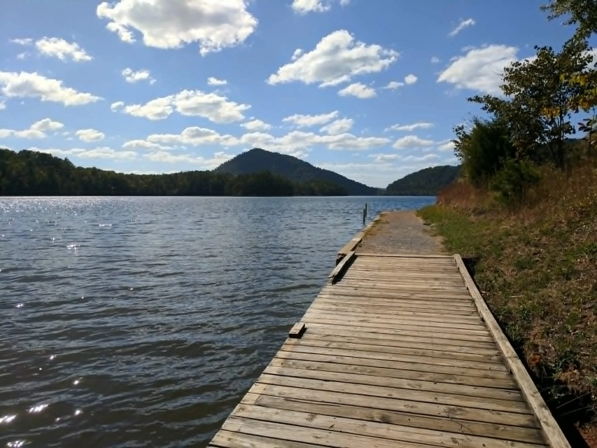 A wooden dock extends over a body of water surrounded by trees and hills under a partly cloudy sky.