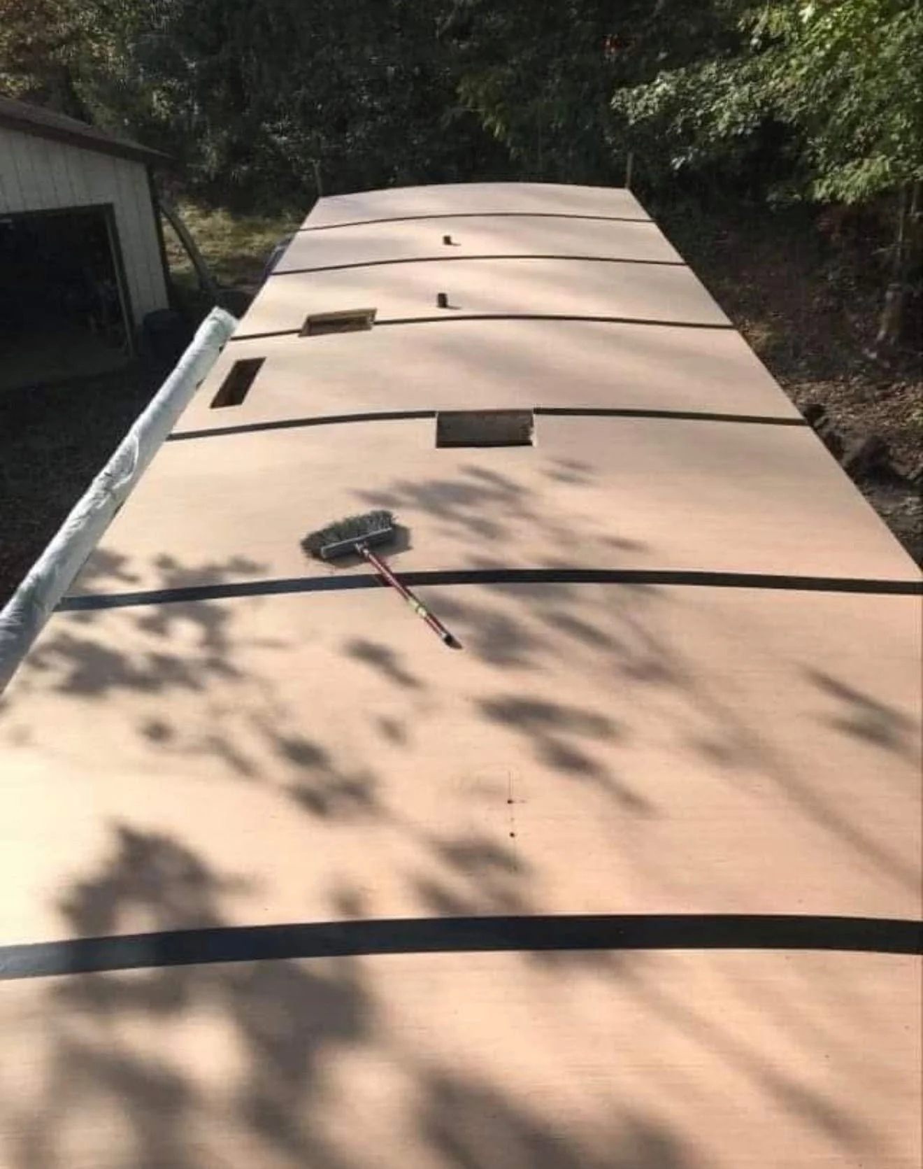 A large flat surface, possibly the top of a boat or table, is being cleaned with a long-handled brush. There is a broom on the surface and a white pipe or pole on the left side. Shadows of trees are cast across the surface, and a small shed or building with an open door is in the background, surrounded by trees and greenery.