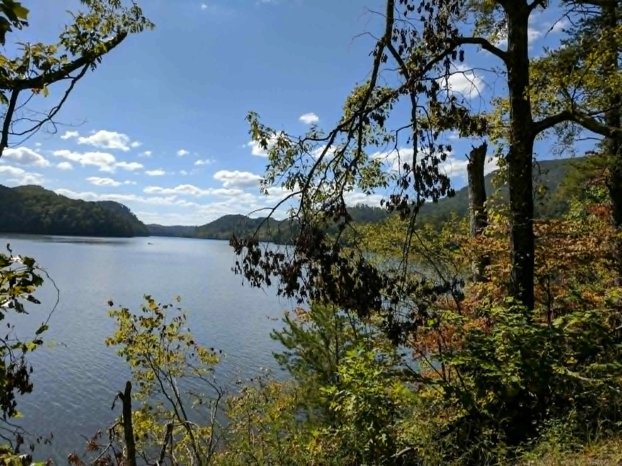 A scenic view of a river under a blue sky with scattered clouds, surrounded by green mountains and trees, some with autumn foliage.