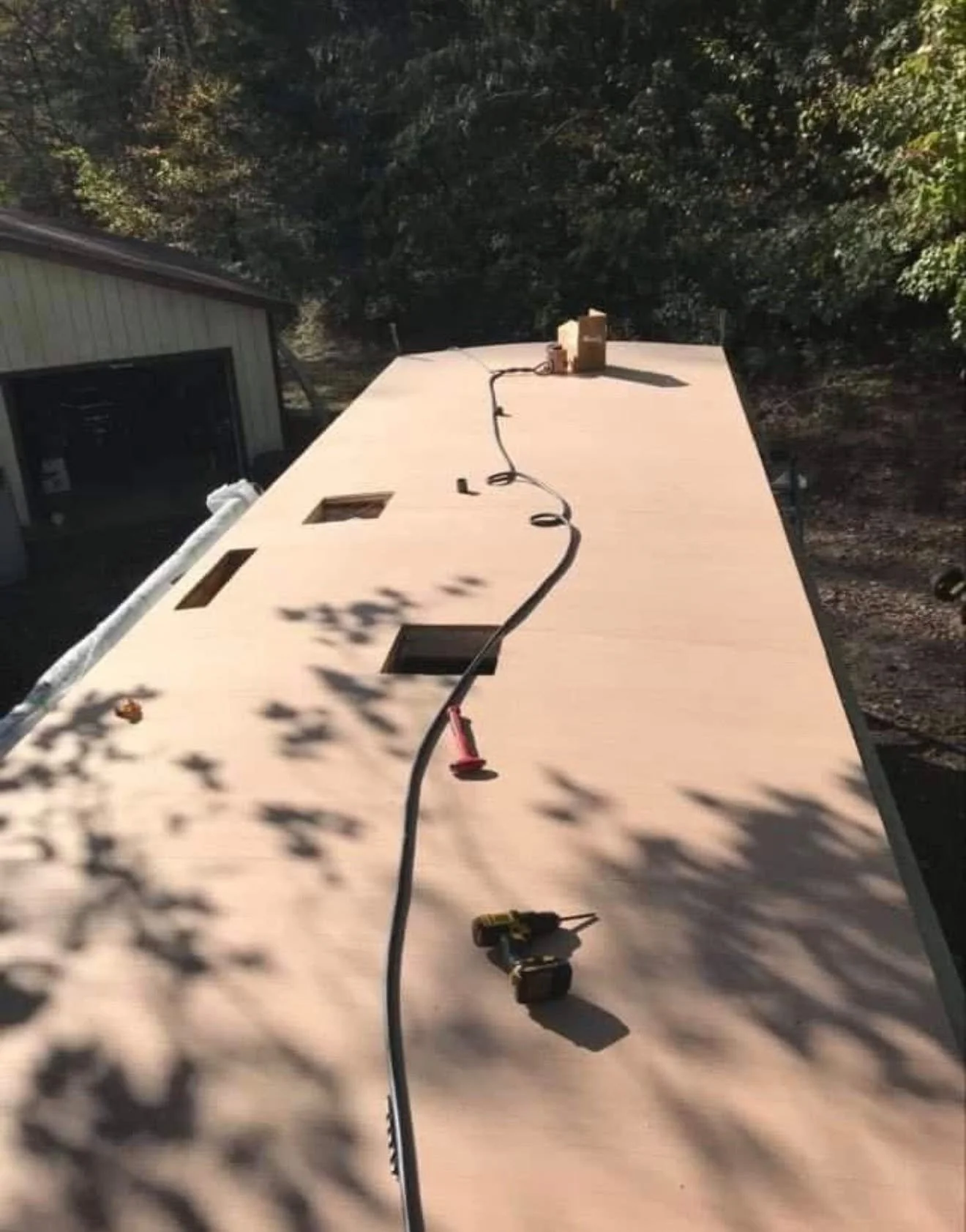 Wooden panel with cutout holes, construction tools, and a cable on it, outdoors in a shaded area with trees.