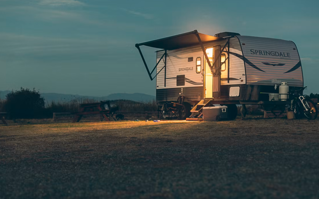 A travel trailer with the brand name 'Springdale' parked in an open field at dusk, with a small outdoor setup including a cooler, propane tank, and a motorcycle nearby.