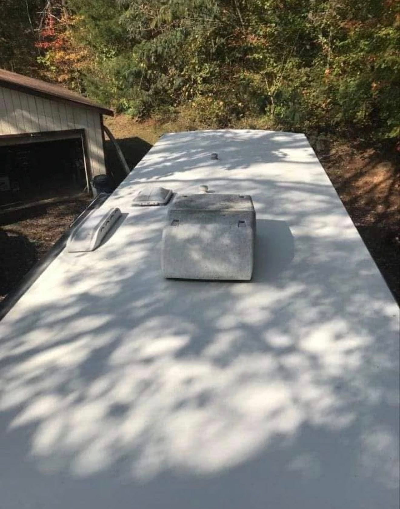 Top view of a white camper van roof with a vent and a small hatch, with trees casting shadows on it, and a neighboring building with an open garage door in the background.