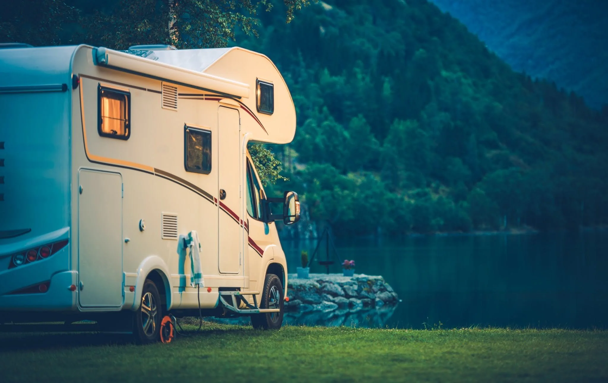 A white camper van parked near a river with a scenic mountain backdrop, camping gear hanging outside, and a small table with flowers nearby.