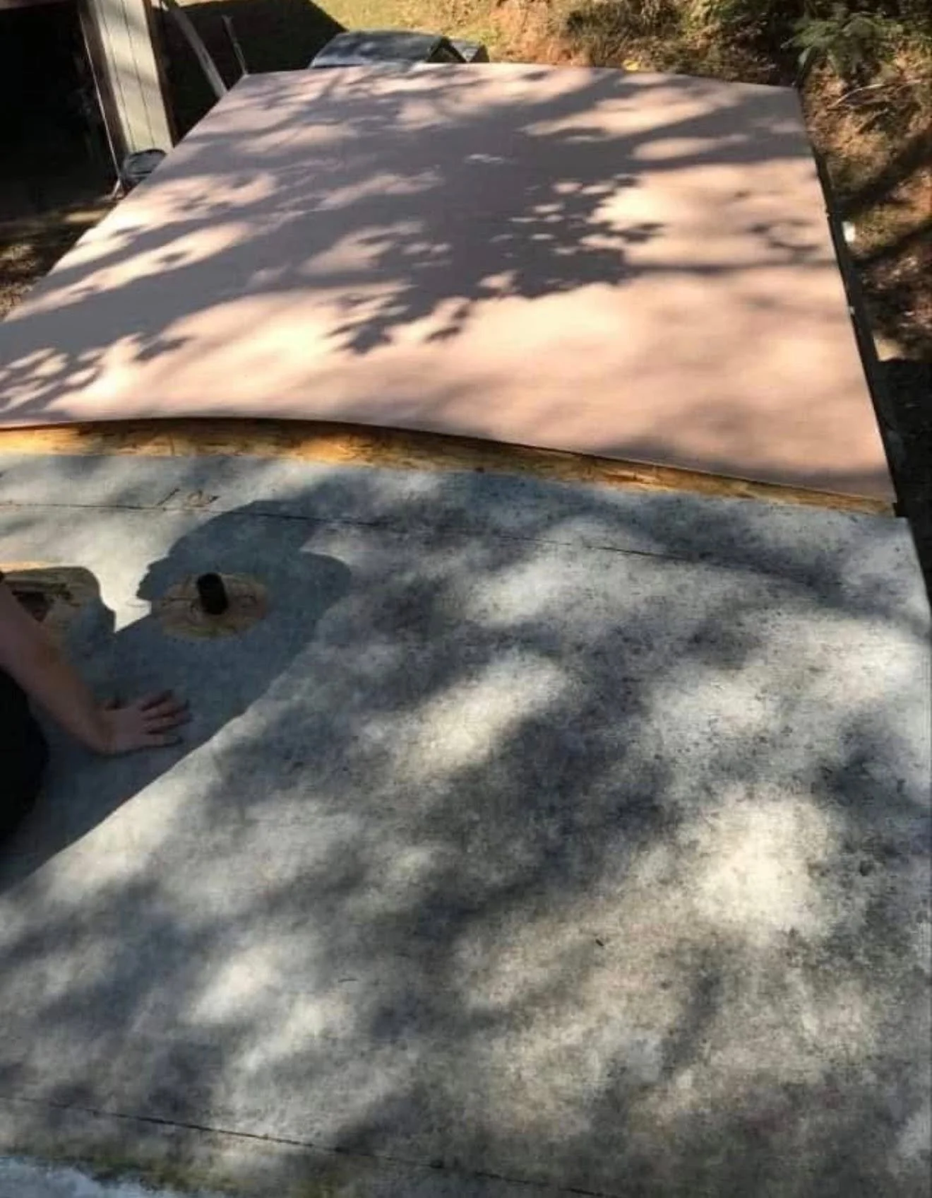The photo shows a concrete surface with tree shadows cast above. In the upper part of the image, there appears to be a large object covered with fabric or tarp, possibly on a flatbed or work surface. To the left, a person's arm and hand are visible resting on the concrete, with a circular object nearby. The scene suggests an outdoor setting with natural light and shadows from the surrounding trees.