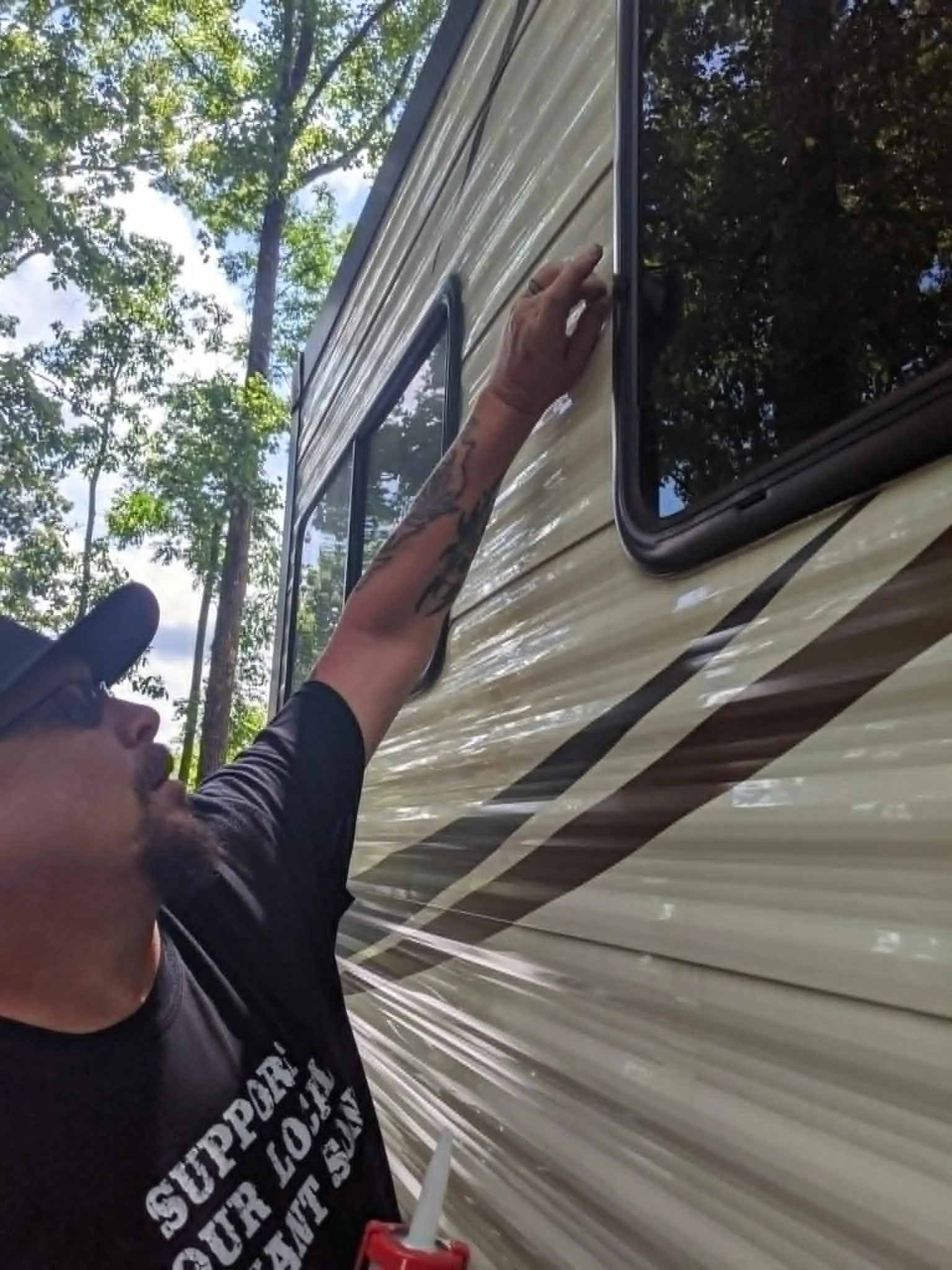 A person using a caulk gun to seal or repair a window in a recreational vehicle (RV) or camper during the daytime surrounded by trees.