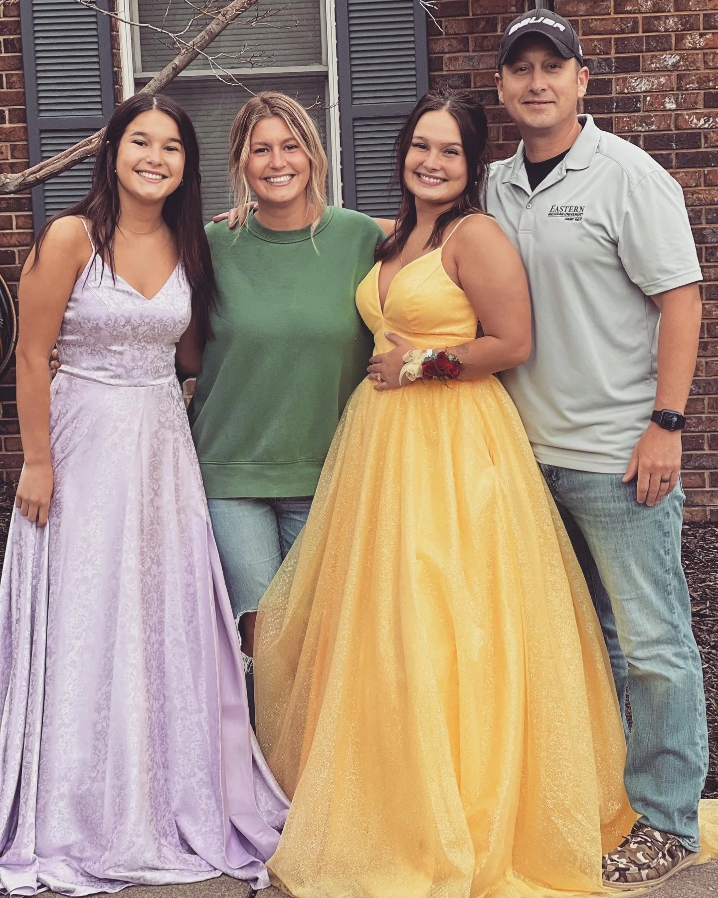 Four people standing outdoors in front of a brick building, smiling, with two women wearing purple and yellow ball gown dresses and a man in casual attire.