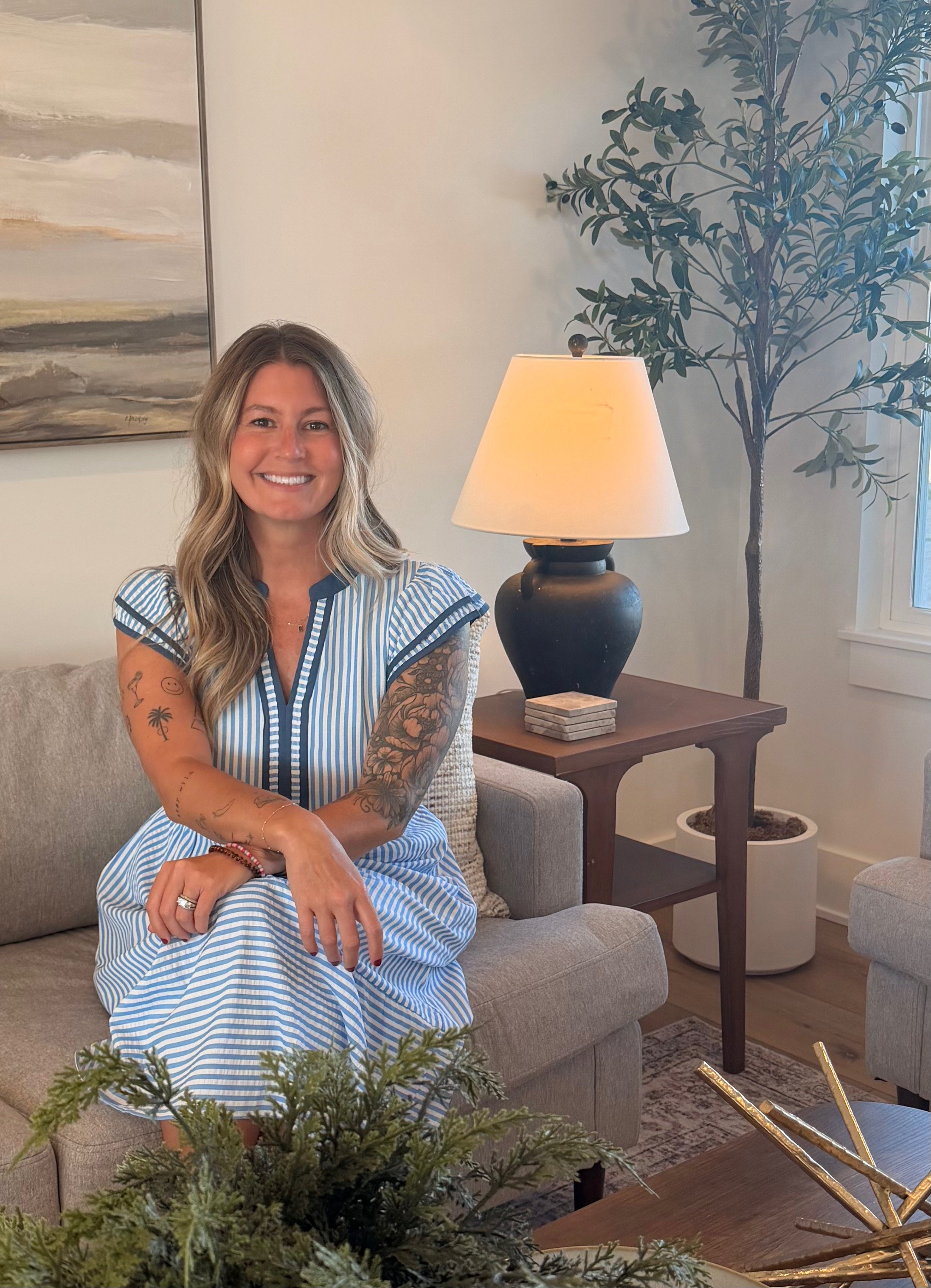 A woman with long, wavy hair and tattoos on her arms, wearing a blue and white striped dress, sitting on a beige couch in a living room. There is a large painting on the wall, a table lamp on a side table, and a tall potted plant in the background.
