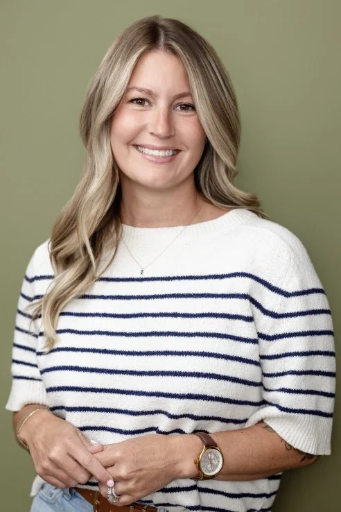 A smiling woman with long blonde hair, wearing a white and navy striped sweater, standing against a green background.