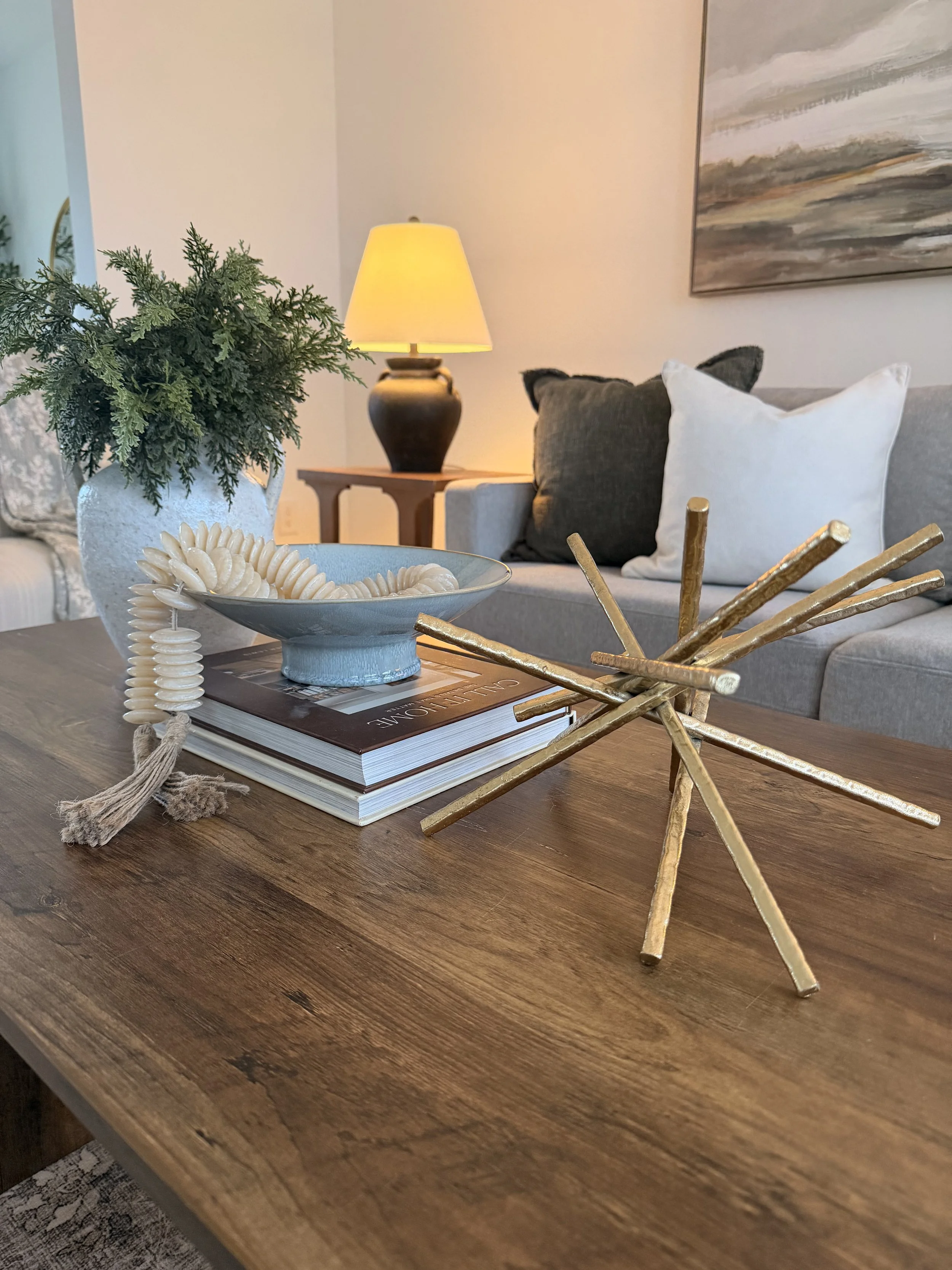 Close-up view of a wooden table with decorative objects including a white bowl with shell-like jewelry, a stack of magazines, a gold starburst ornament, a green plant in a white textured pot, a beige table lamp, and a background scene with a gray sof