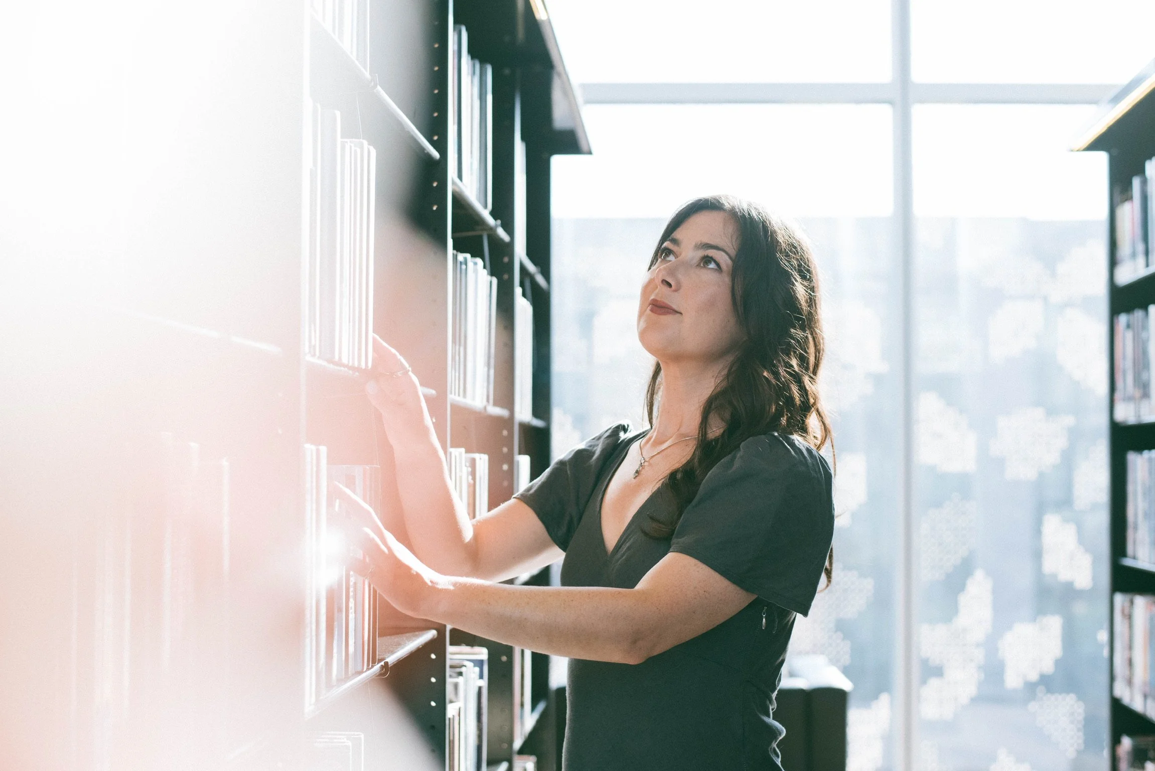A woman with dark wavy hair wearing a dark gray top is reaching for a book on a bookshelf in a well-lit library with large windows in the background.
