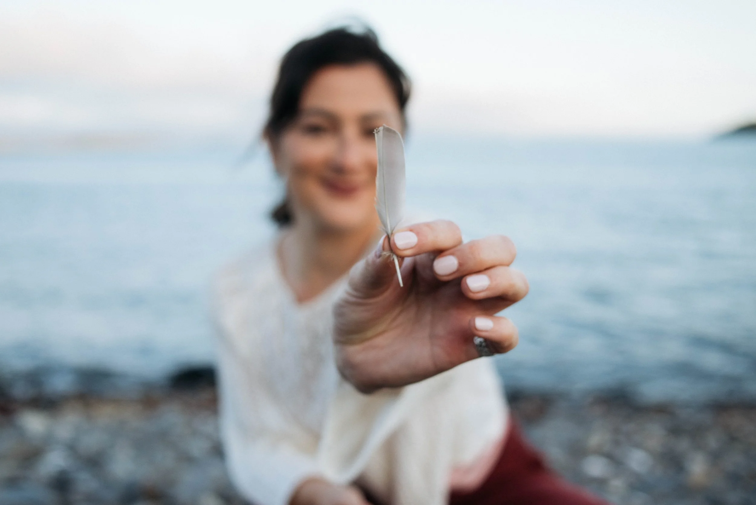 A woman with dark hair holding a white feather feather close to the camera, smiling in the background near water.
