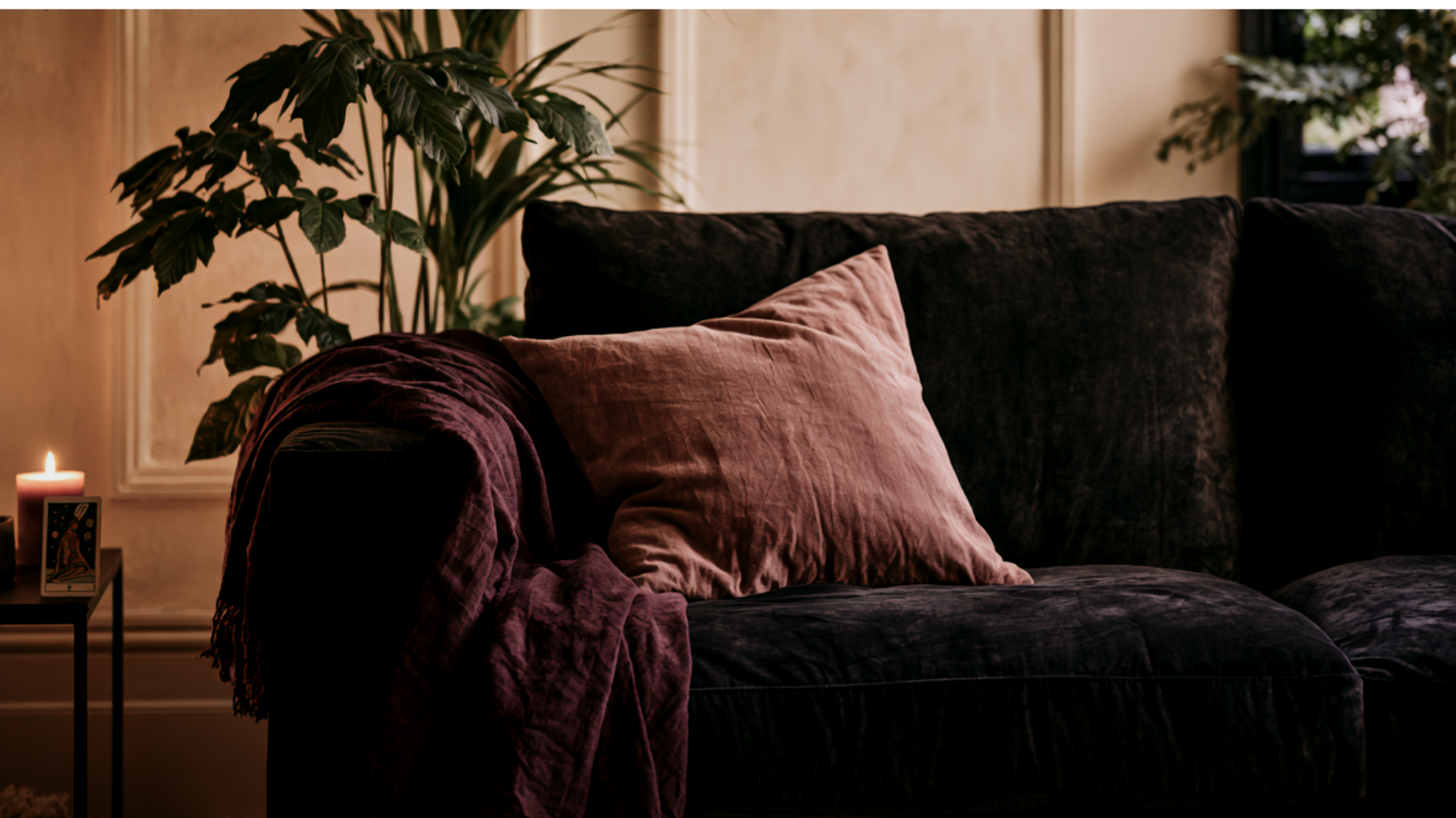 A cozy living room with a dark velvet sofa, pink pillow, and purple throw blanket. There is a green indoor plant, a candle, and a small religious card on a side table. Soft ambient lighting creates a warm atmosphere.