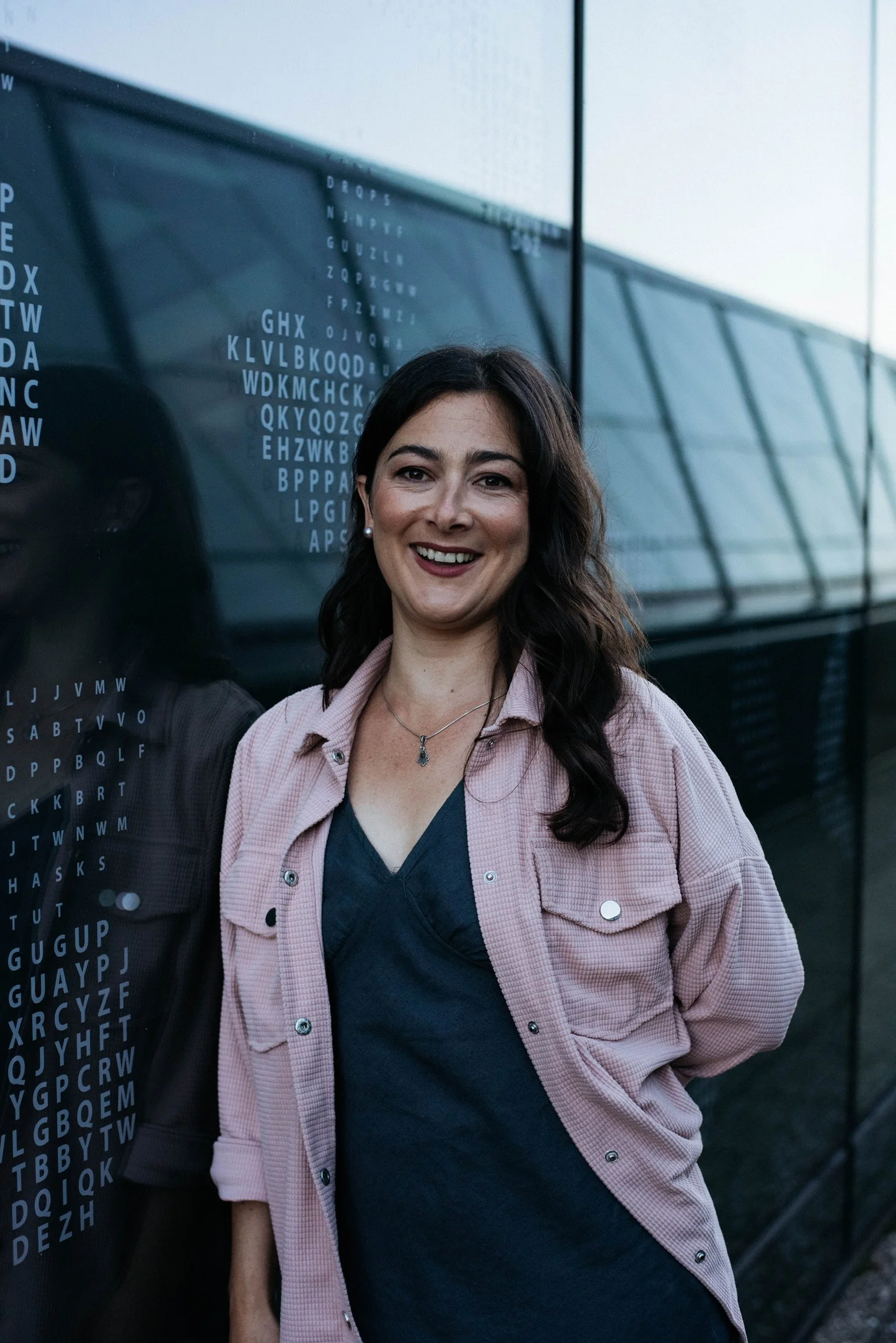 A smiling woman with dark hair and pearl earrings standing in front of a reflective glass wall with alphabet letters and eye charts. She wears a dark top and a pink jacket, with her hands behind her back.