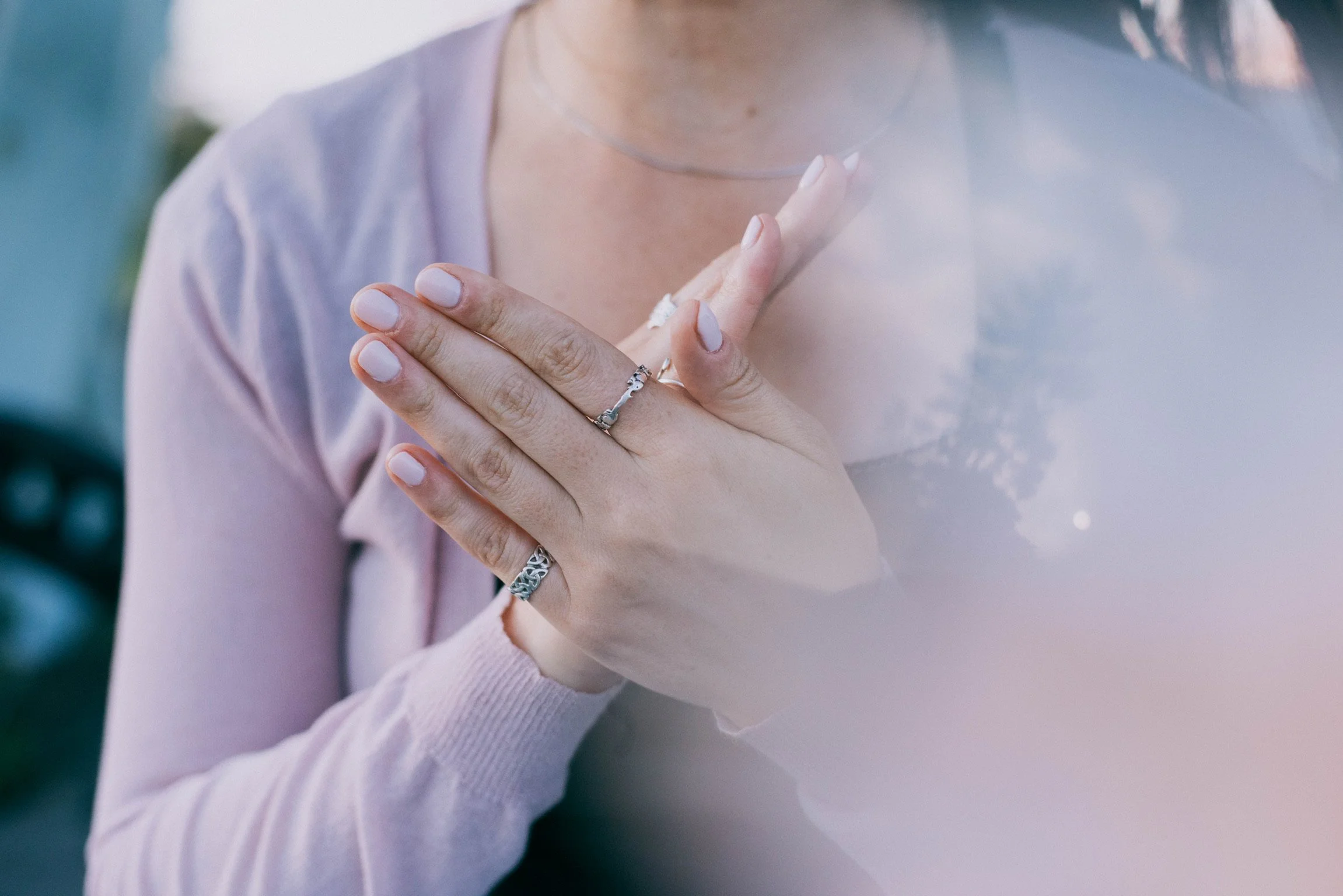Close-up of a woman placing her hand on her chest, wearing rings and a light pink sweater, with soft natural lighting.