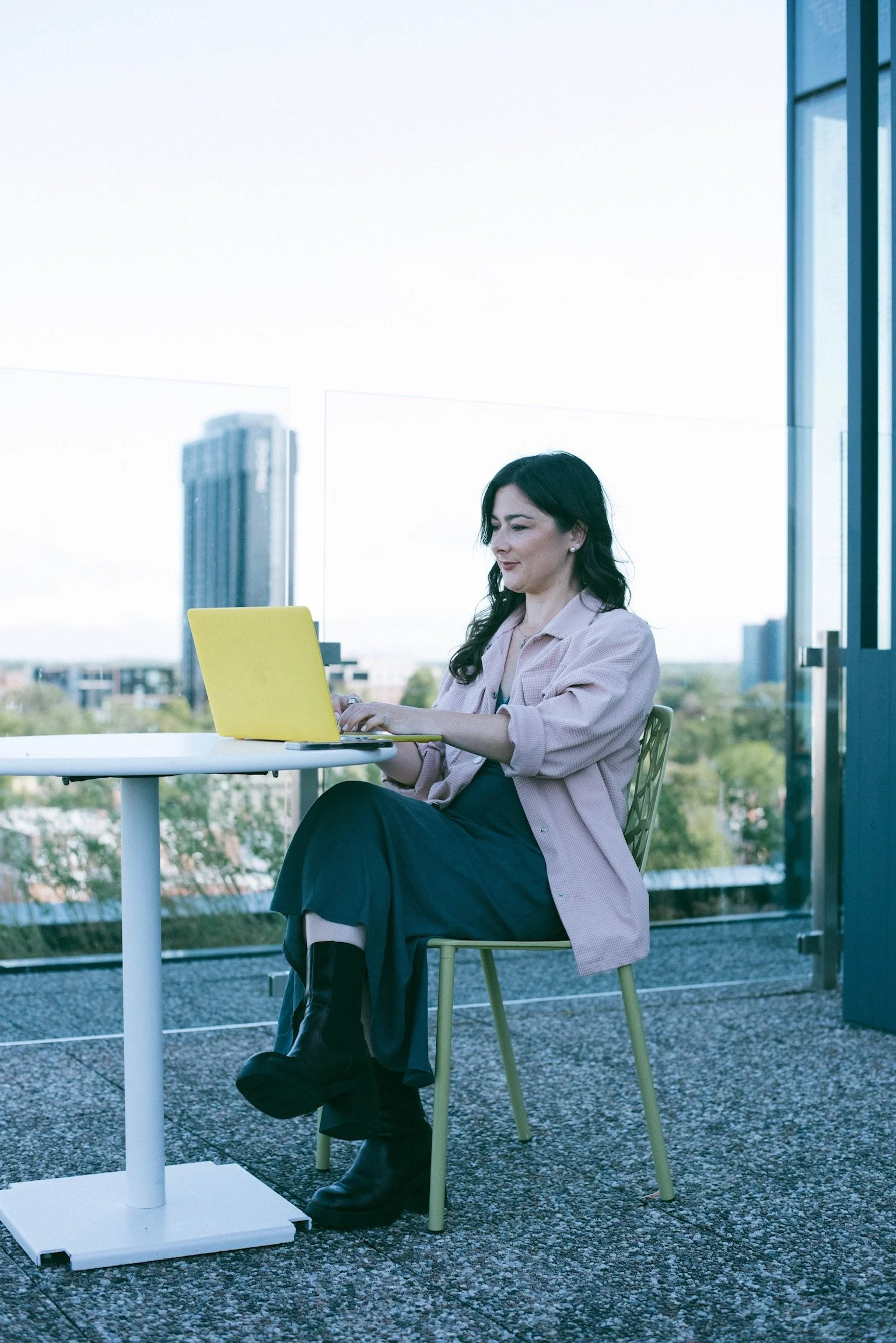 A woman with dark hair wearing a light pink blazer and black boots is sitting at a white outdoor table with a yellow laptop, indoors near large windows showing a cityscape with tall buildings.