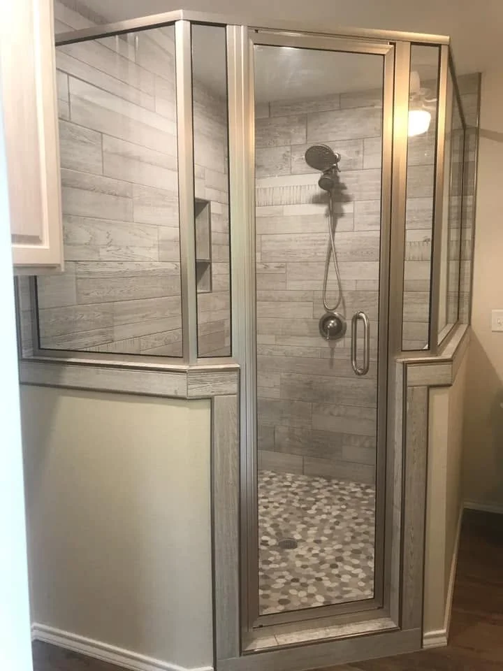 A glass shower enclosure with a metal frame, tan and gray wood-like tile walls, and a pebble stone floor.