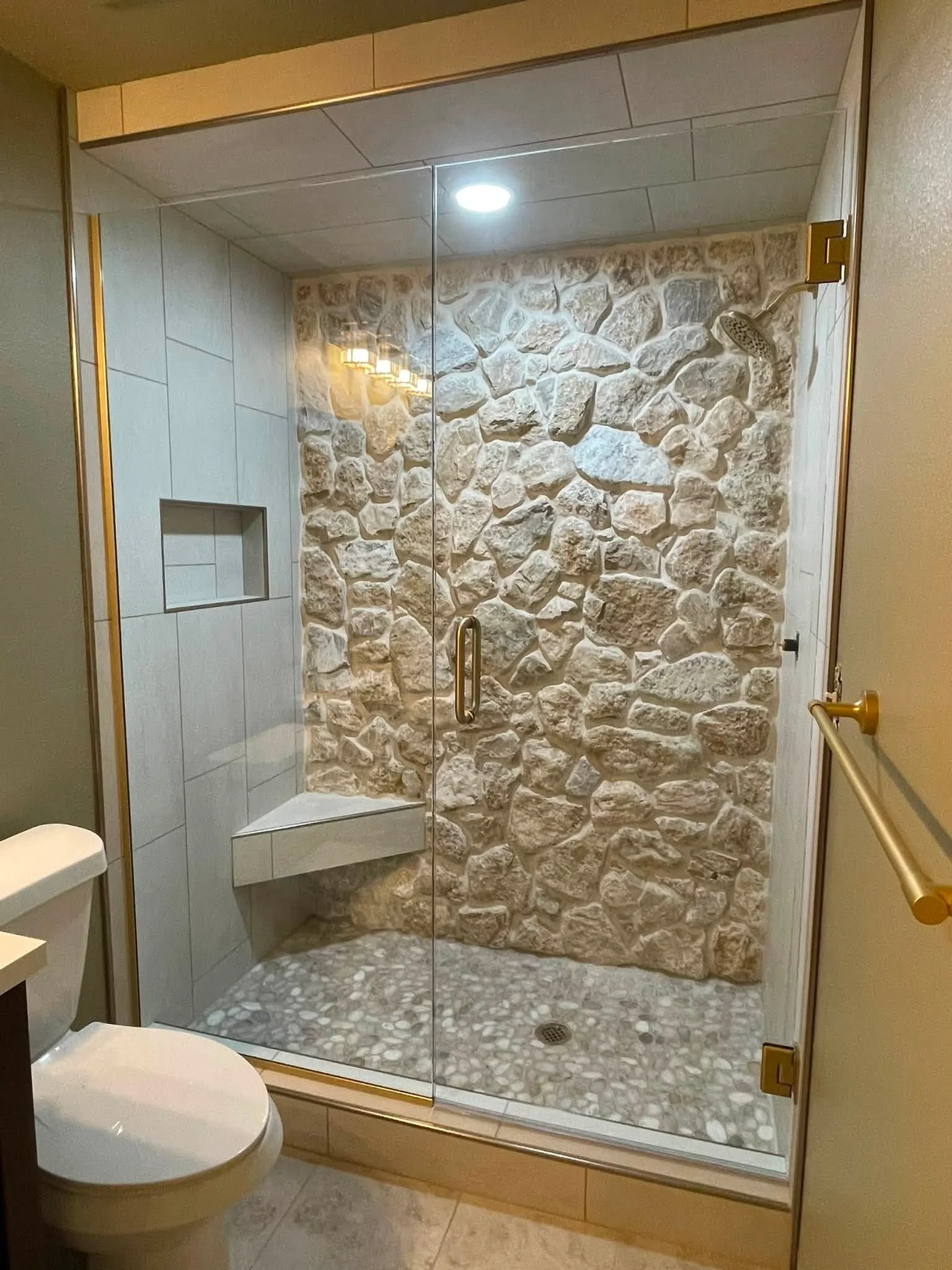 A bathroom shower with a large glass door, stone wall interior, and pebble floor, adjacent to a toilet.