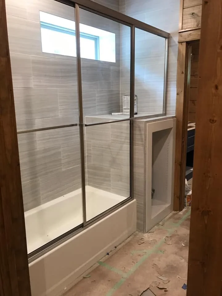 Bathroom with a shower area enclosed by glass doors, gray tiles on the walls, a small window above, and a white bathtub base. There is a built-in niche with shelves in the wall, and unfinished flooring.