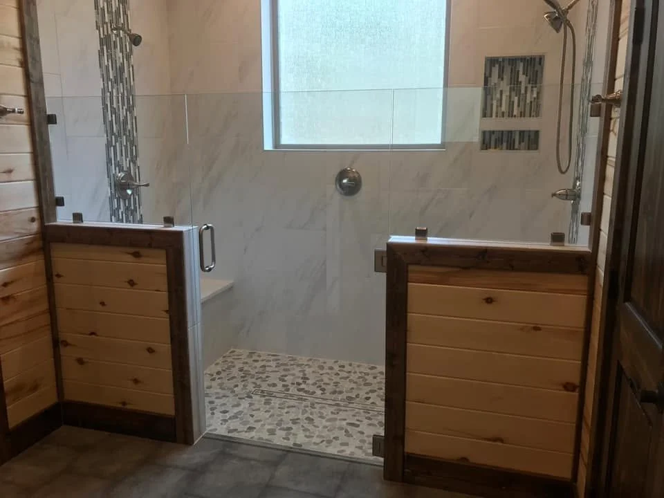 A walk-in shower with a glass door, tiled walls, a window for natural light, and pebble stone flooring, surrounded by wood-paneled walls.