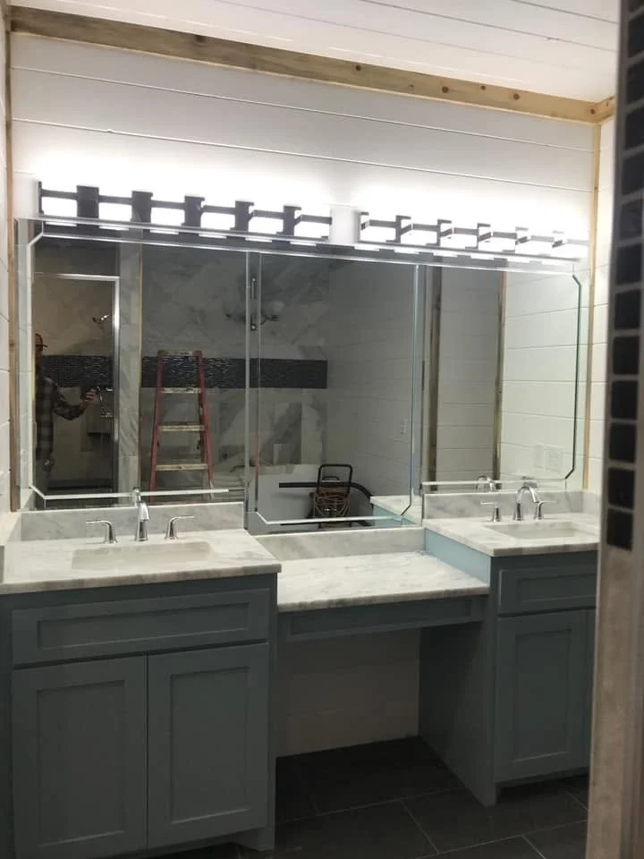 Double bathroom vanity with marble countertops, two sinks, and a large mirror with lights above. A reflection of a person taking the photo, a ladder, and other bathroom elements are visible in the mirror.