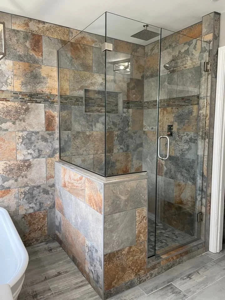 A modern glass shower with slate tile walls, a built-in tile niche, and a rain showerhead. The shower is enclosed with clear glass panels and a glass door, with a tiled half-wall at the entrance.