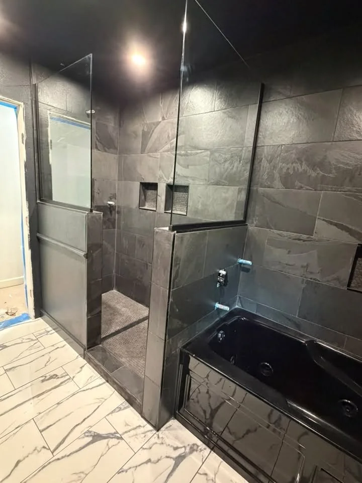Bathroom with a black bathtub, gray tiled walls, a walk-in shower area with glass partition, and cream-colored marble floor tiles.