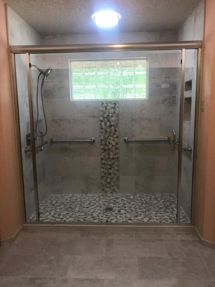 A walk-in shower with glass doors, a handheld showerhead on a hose, and a small window letting in natural light. The shower has beige and brown tile with a vertical strip of pebble mosaic tiles. There are two metal grab bars inside.