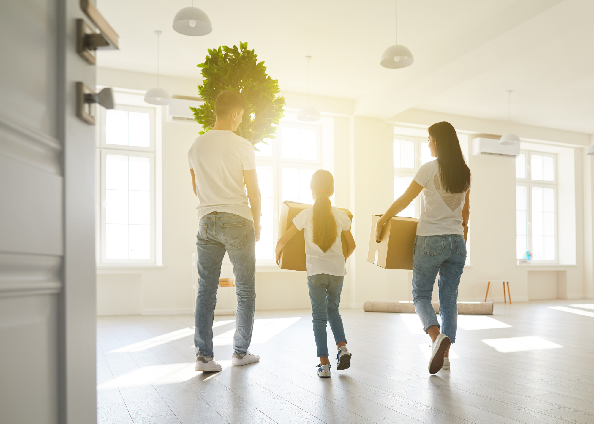 A family moving into a new home, with the parents and a girl carrying cardboard boxes, walking through a bright, spacious living room.
