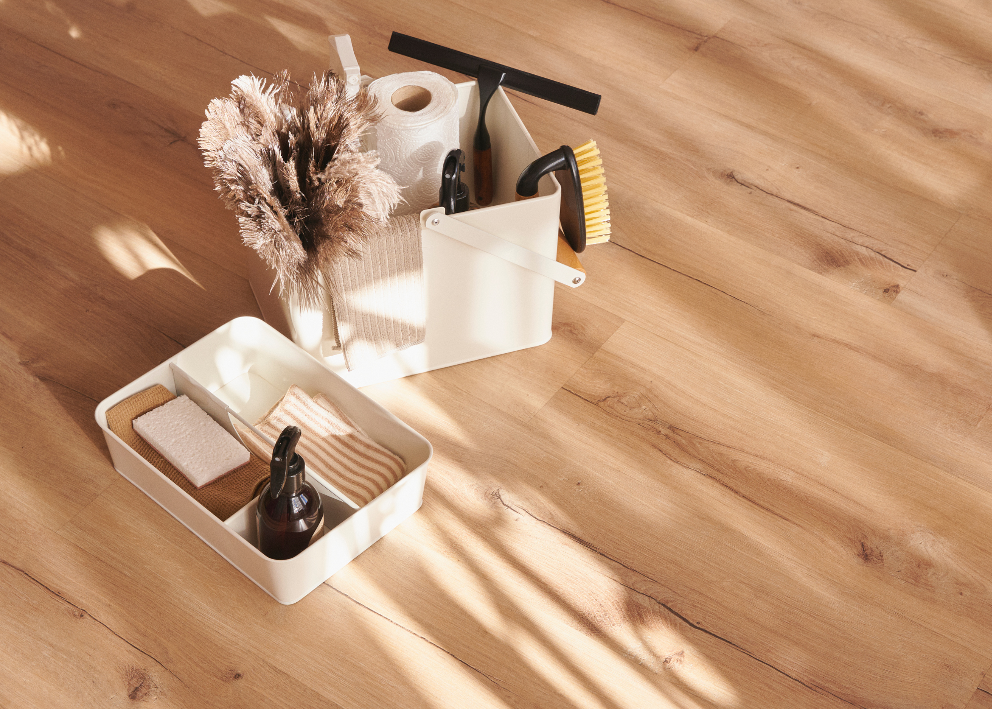 A white storage bin on a wooden surface containing a roll of paper towels, dried flowers, a small beige cloth, and cleaning tools such as brushes and a spray bottle, with sunlight casting shadows.