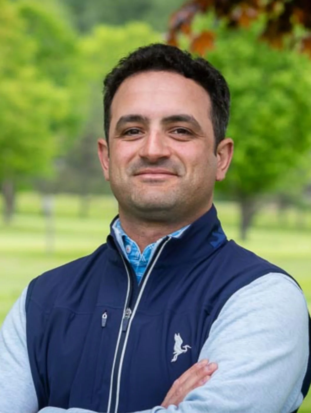 A man with dark hair smiling outdoors with green trees in the background wearing a navy vest over a light gray long-sleeve shirt.
