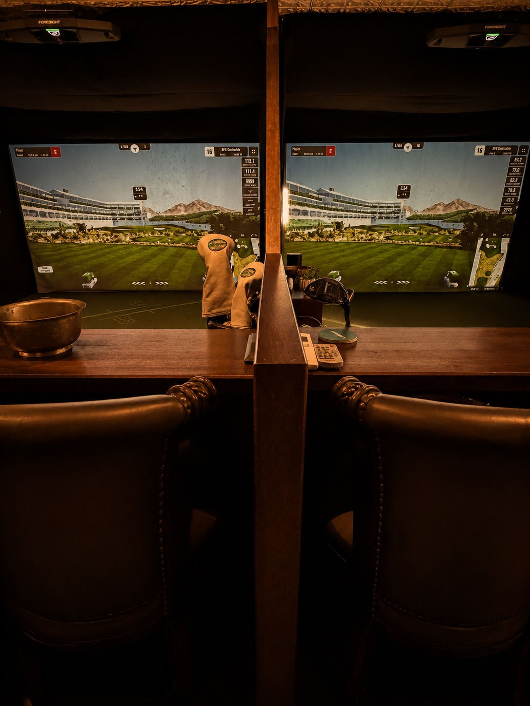 Indoor golf simulation room with two monitors displaying a golf course, golf clubs, gloves, and related accessories on the table in front of chairs.