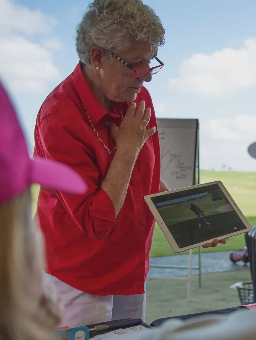 An elderly woman with curly gray hair, red glasses, and a red shirt is holding a tablet displaying a golf swing. She is inside a tent, with a golf course visible in the background. She has a hand on her chest and is looking at the tablet.