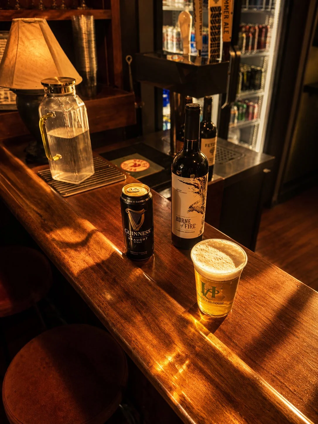 A bar with a wooden counter, featuring a pint of beer, a can of Guinness, a bottle of wine labeled 'Borne of Fire,' and a glass water dispenser with a yellow handle. The background includes liquor bottles and a cigar box.