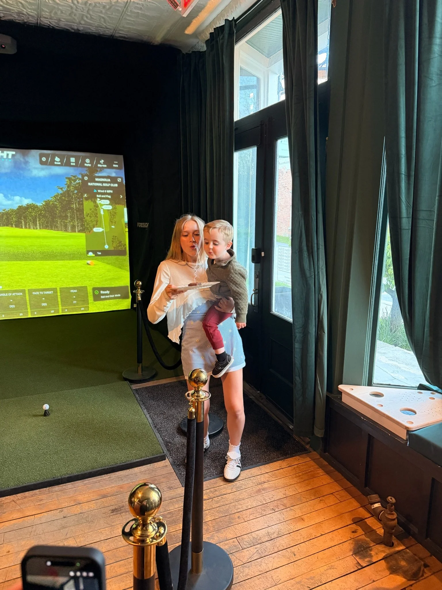 A woman holding a young boy while standing in an indoor golf simulation area. There is a large screen showing a golf course with a tee, and the area is cordoned off with black and gold stanchions. The room has wooden flooring, large windows with dark curtains, and some outdoor scenery visible through the glass.
