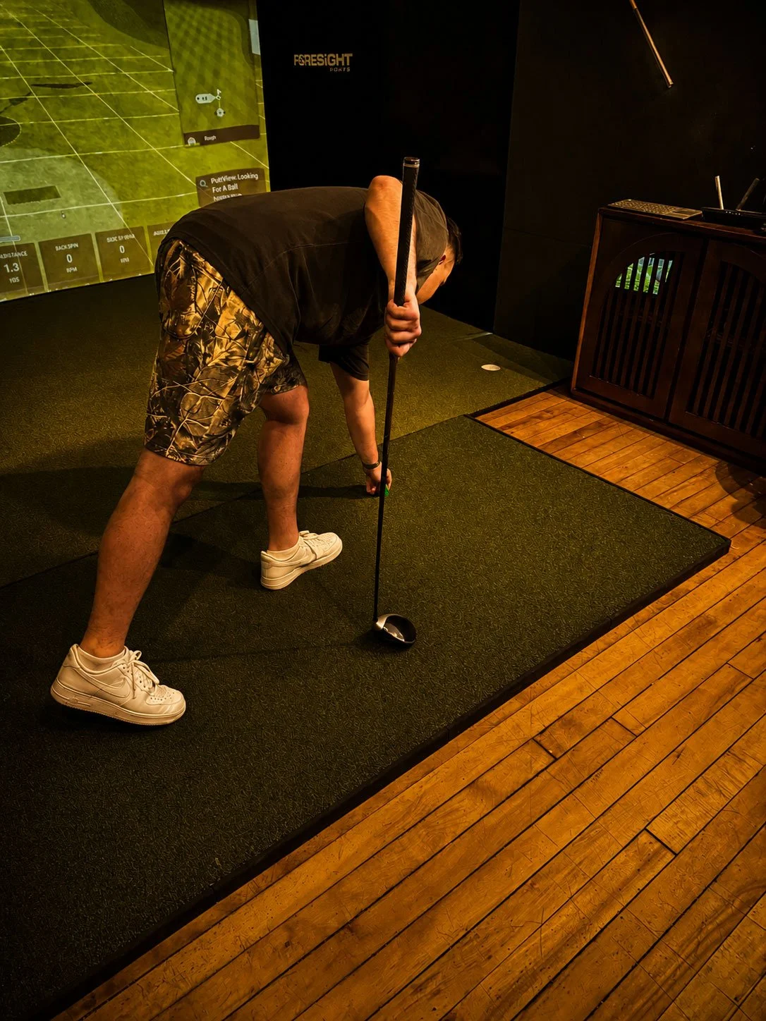 Man practicing his golf swing indoors, bending over with a golf club in hand, wearing camouflage shorts, a black t-shirt, white sneakers, and a cap, on a small carpeted area.