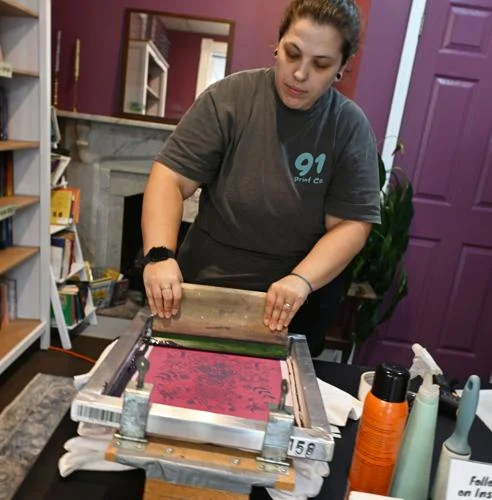 A person operating a printing press in a room with bookshelves and purple walls.