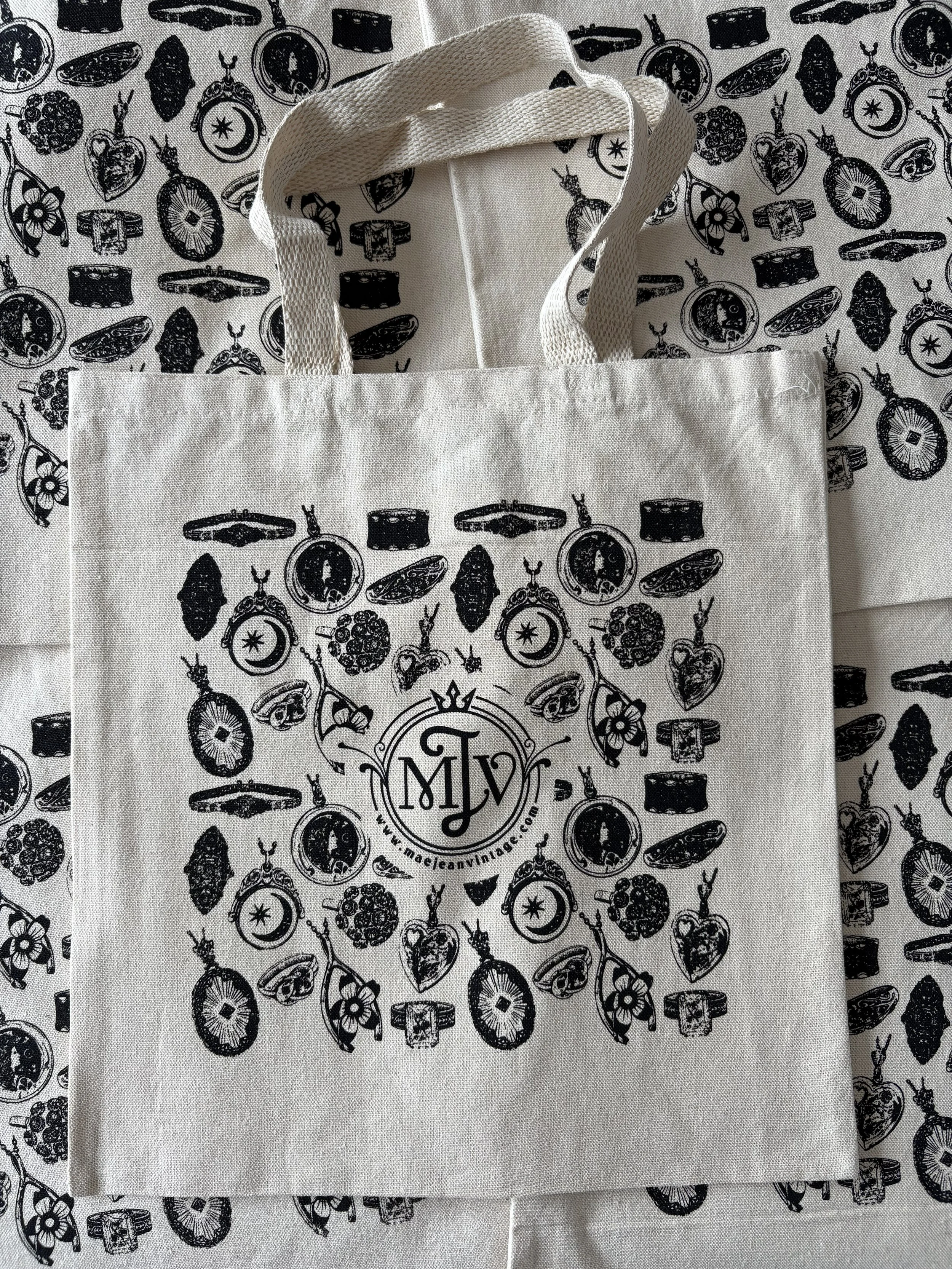 A natural tote bag with a black printed pattern of vintage jewelry and a circular logo that says 'Maejean Vintage'