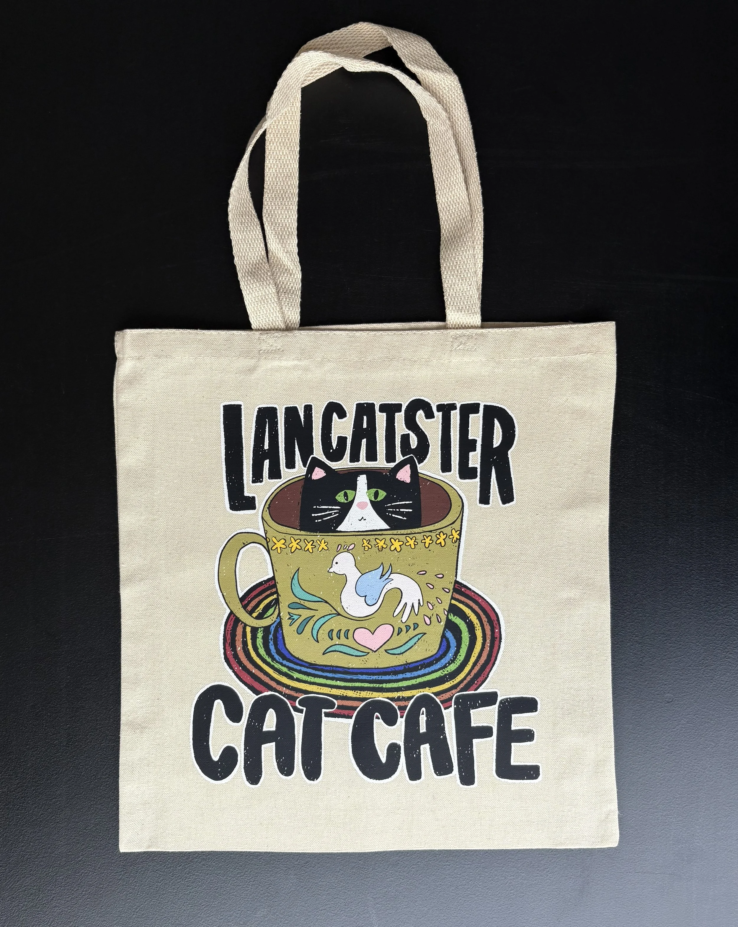 A natural colored tote bag with the words "Lancatster Cat Cafe" printed on it, featuring a colorful illustration of a black and white cat inside a coffee cup with a rainbow swirl pattern.